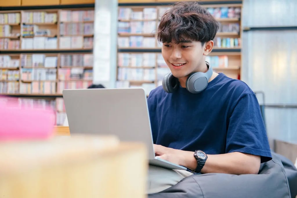Private online homeschool student smiling while studying on a laptop in a library setting with bookshelves in the background.