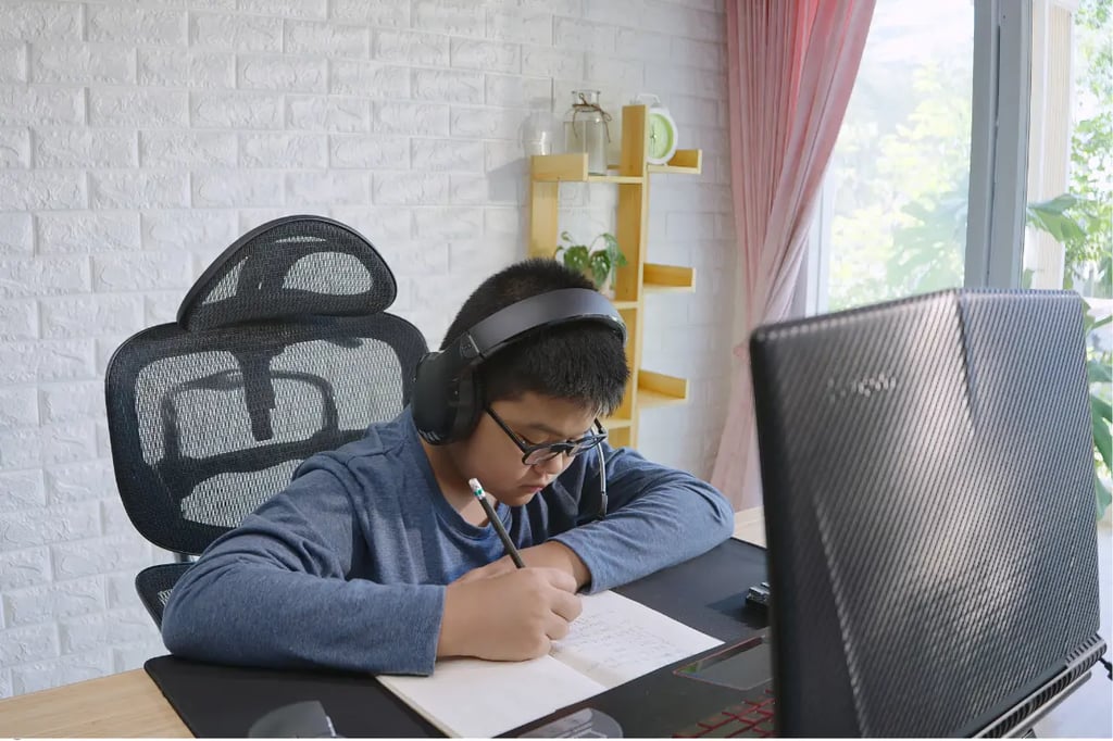 Accredited homeschool student wearing headphones and writing in a notebook while studying online at a desk with a laptop.