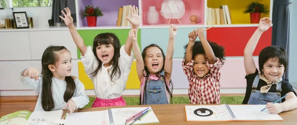 Experiential learning shown by enthusiastic young children engaging in creative classroom activities with raised hands and joyful expressions.