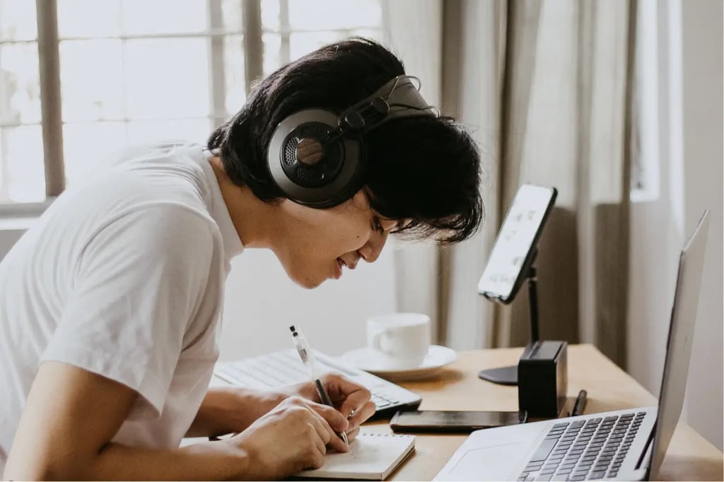 Project based learning student focused on writing notes while wearing headphones and working at a laptop