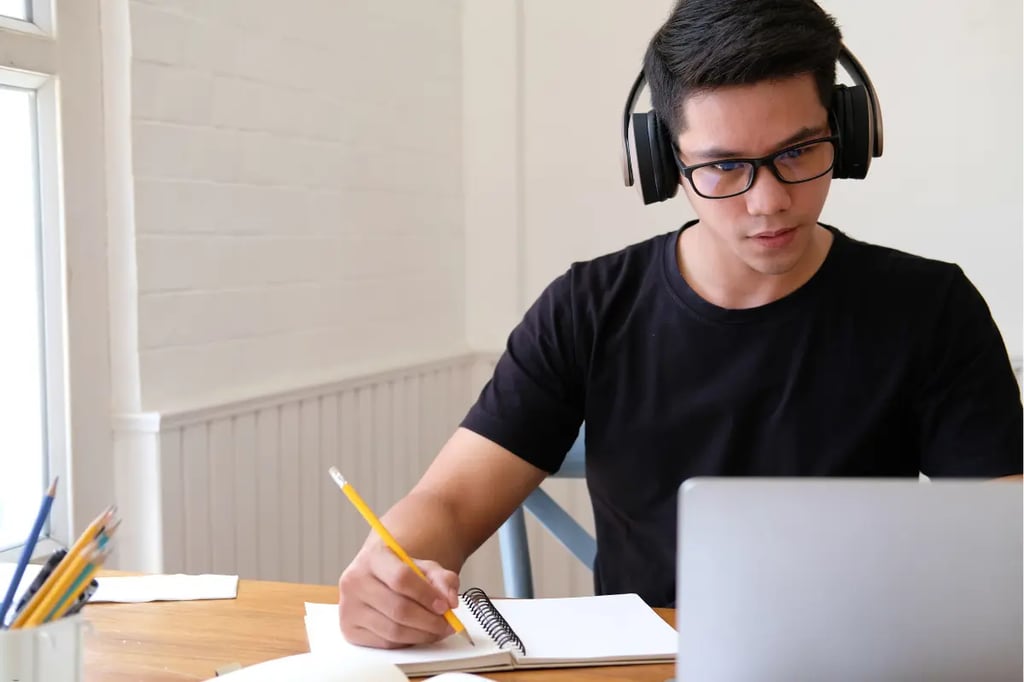 Private online homeschool student wearing headphones, taking notes while studying on a laptop at a wooden desk