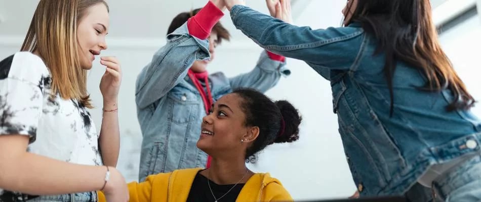 Online high school hobbies depicted by a group of happy teens interacting and high-fiving in a collaborative indoor setting.