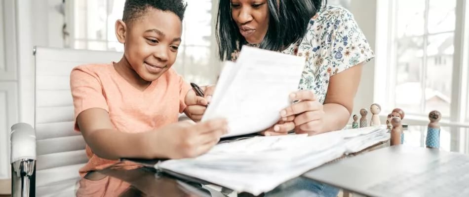 Online homeschooling experience in middle school shown by a supportive parent helping a smiling student with printed assignments.
