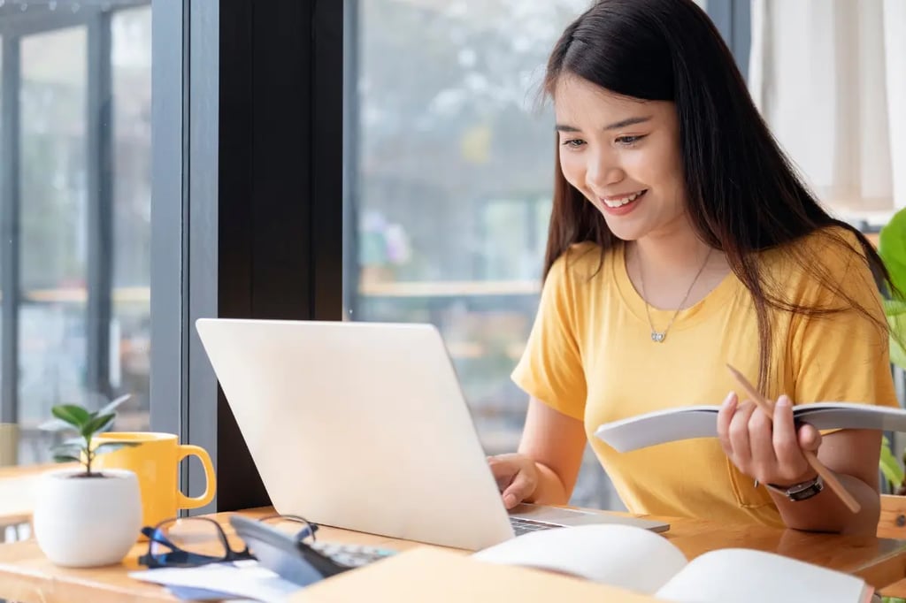 Student engaged in private online homeschool studies, smiling while using a laptop and holding a notebook at a sunny desk