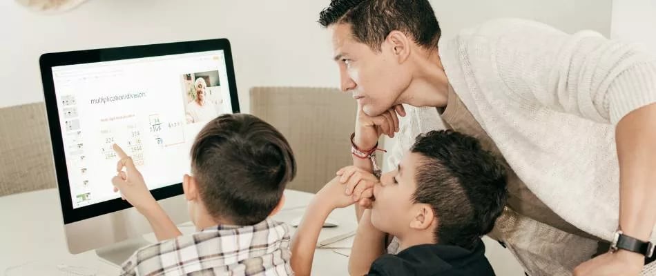 Online homeschooling illustrated by a parent guiding two young children through a digital learning activity on a desktop computer.