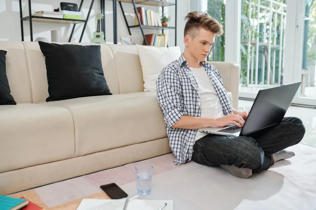 A young man sits on the floor using his laptop, participating in private online homeschooling from his living room