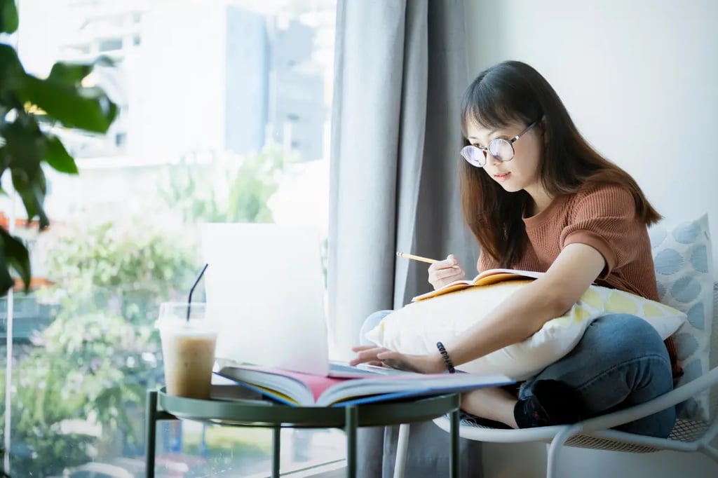 Young woman studying with a notebook and laptop near a window, with an iced coffee on a table nearby.
