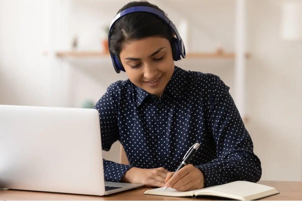 Online learning student wearing headphones, taking notes while studying on a laptop at home