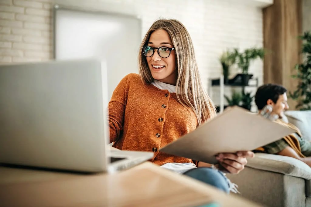A woman wearing glasses reviews documents and works on personalized lesson plans using a laptop in a cozy setting