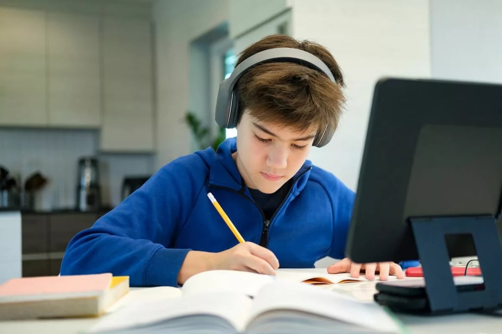 Experiential learning shown by a student deeply engaged in writing while attending an online class with headphones and books.