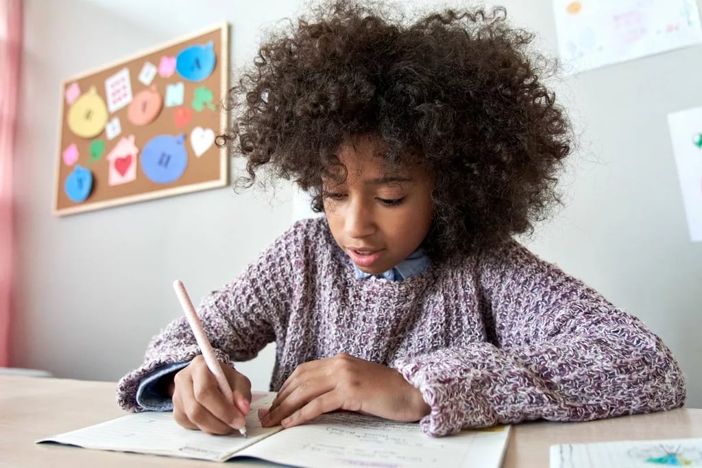 Online homeschool captured by a focused young girl writing in a notebook at home with colorful art on the wall.
