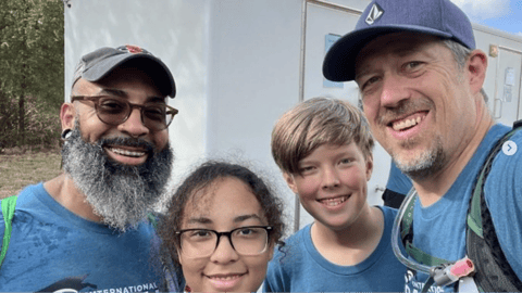 A group of four smiling people wearing matching blue shirts take a selfie outdoors during a community or volunteer event.