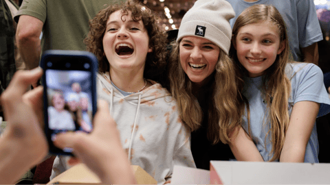 Three smiling friends pose for a photo while someone captures the moment on a smartphone at a joyful gathering.