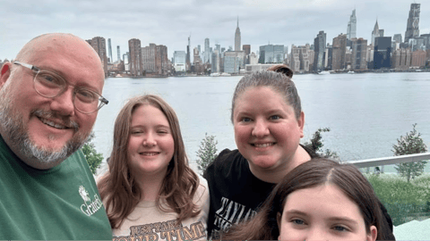 A smiling family of four takes a selfie with the New York City skyline in the background across the water.