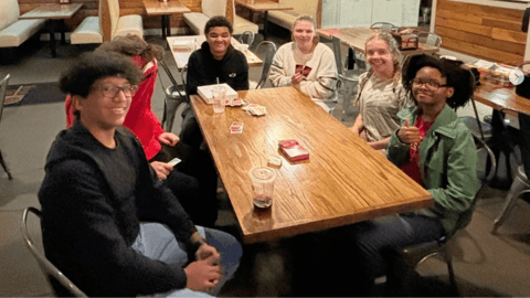 A group of friends sits around a wooden table at a casual restaurant, smiling and enjoying conversation.