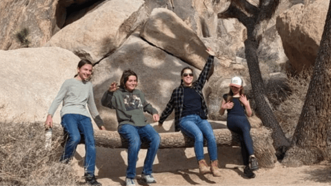A group of four friends smiles and poses while sitting on a fallen tree in a rocky desert landscape.