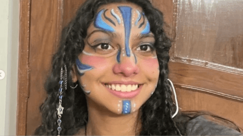 A smiling young woman with curly hair wears intricate blue and white face paint, posing in front of a wooden door.