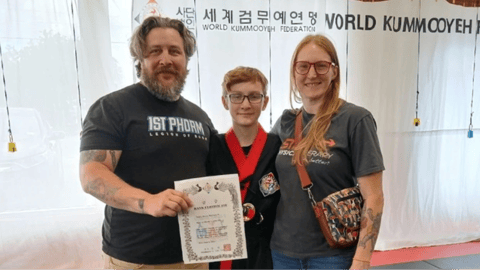 A young martial artist in a black uniform with a red collar stands proudly with their parents, holding a certificate at a World Kummooyeh event.