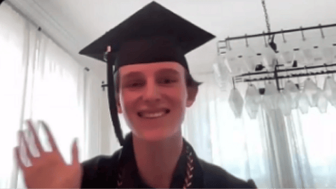 Private online high school graduate wearing a cap and gown smiles and waves during a virtual graduation ceremony at home.