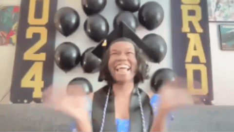 Private high school graduate wearing a cap and gown celebrates joyfully at home with graduation decorations and black balloons.