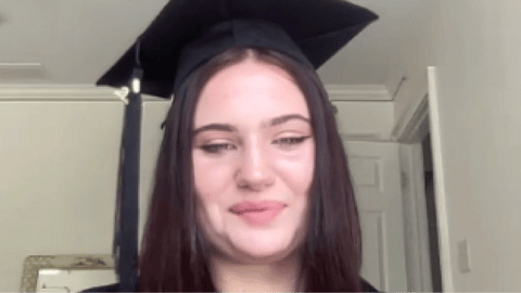 Private online high school graduate wearing a cap and gown smiles during a virtual graduation ceremony at home.