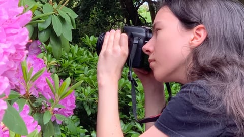 A young woman with dark hair and a black shirt photographs pink flowers with a DSLR camera in a lush garden.