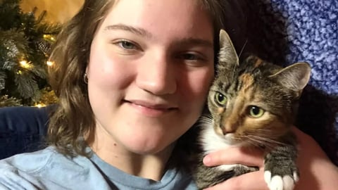 A young woman smiling while holding a tabby cat close to her face, with a Christmas tree in the background.