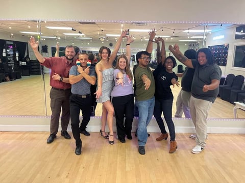 A group of people posing excitedly in a dance studio with mirrors and wooden flooring.