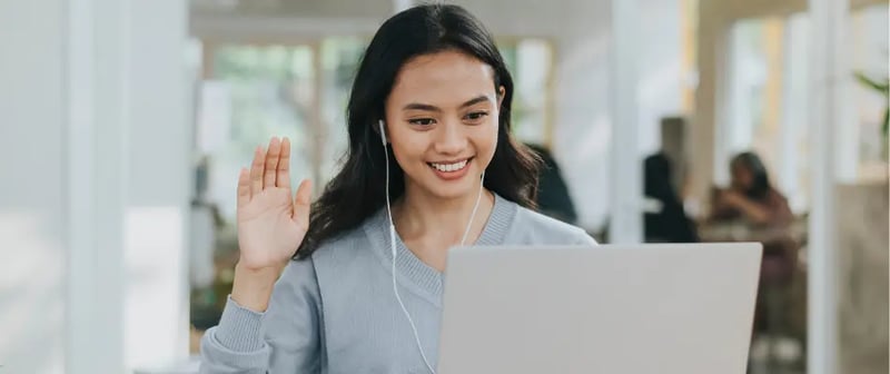Online highschool student smiling and waving during a virtual class on a laptop with earphones in a bright room.