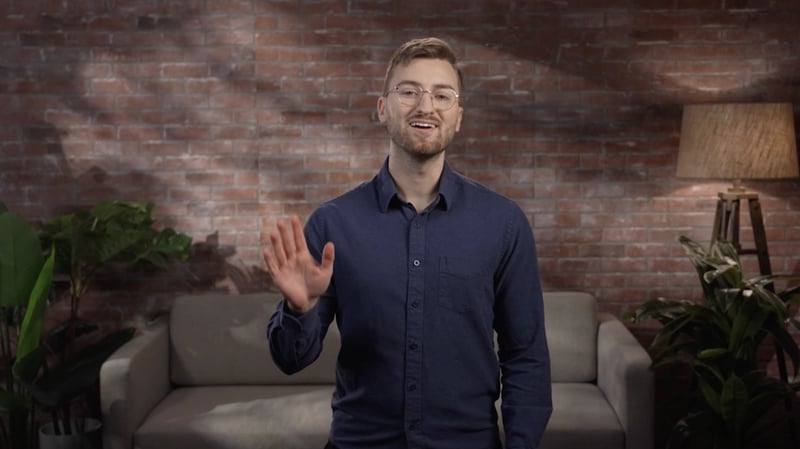Private online school CEO in a navy blue shirt smiles and waves while speaking in a warmly lit, modern office setting.