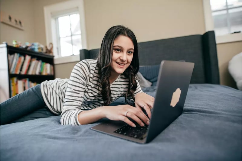 Homeschooling benefits include flexible learning as shown by a smiling girl using a laptop on her bed at home.