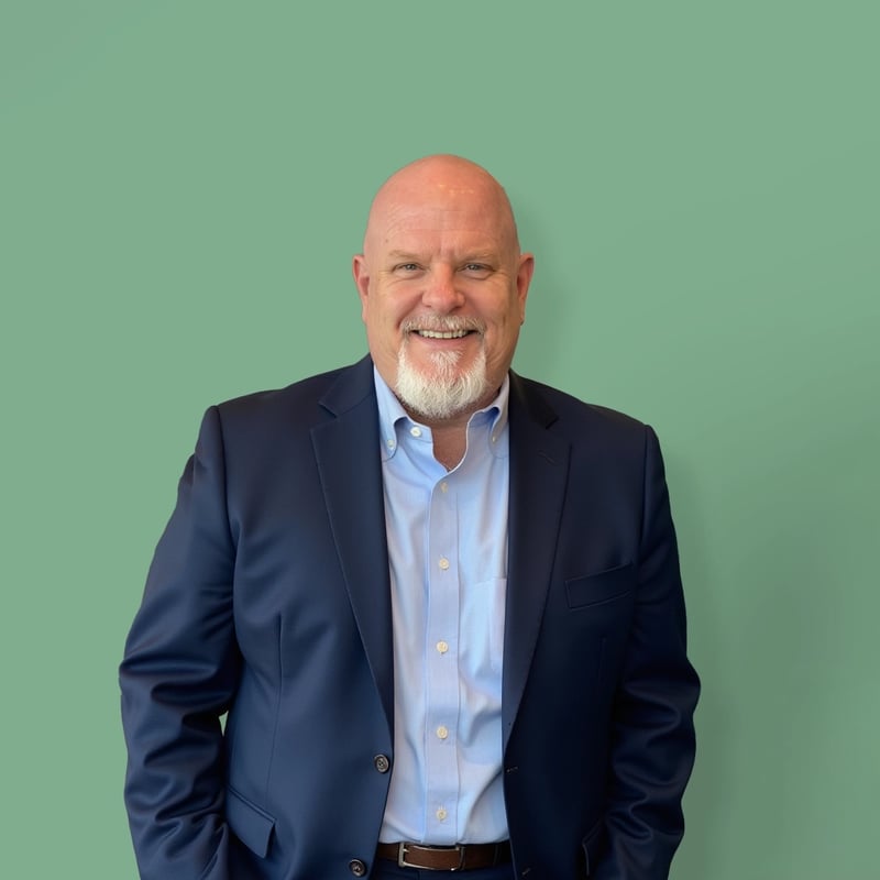 Private online high school director in a navy blue suit and light blue shirt smiles while posing against a green background.