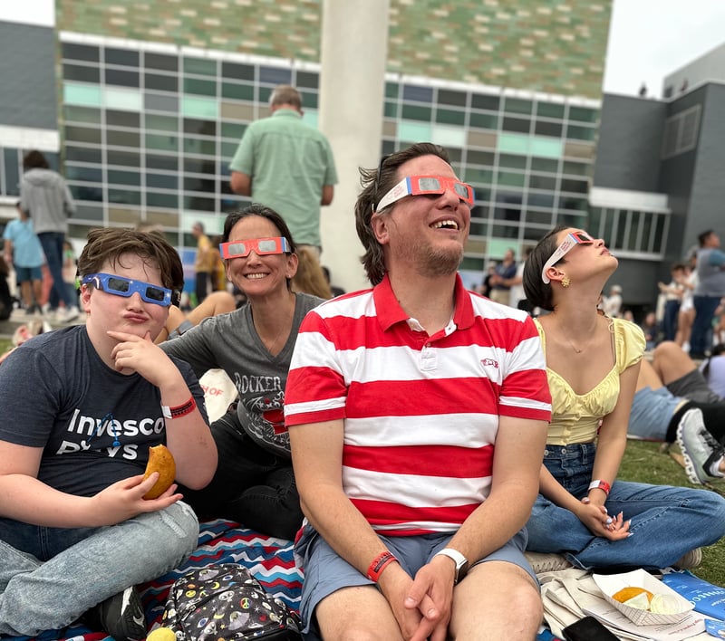 A group of people wearing eclipse glasses sit on a blanket outdoors, looking up at the sky during a solar eclipse event.