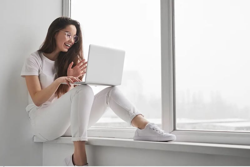 Project based highschool student smiling and waving during an online session while sitting by a large window with a laptop