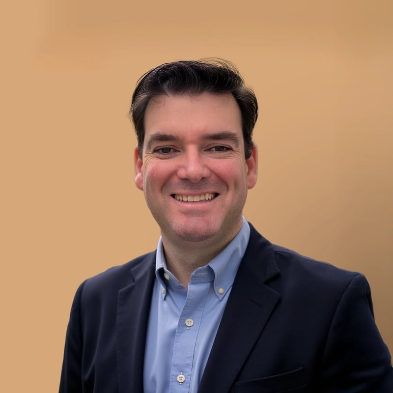 A smiling man in a dark suit and light blue shirt poses against a solid beige background