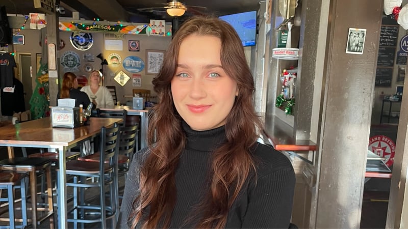 A young woman with long brown hair and blue eyes wearing a black turtleneck smiles while sitting in a casual restaurant.