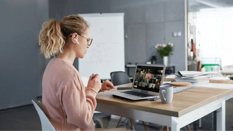 Private online high school student wearing glasses and a pink sweater attends a virtual class on a laptop in a modern study space.