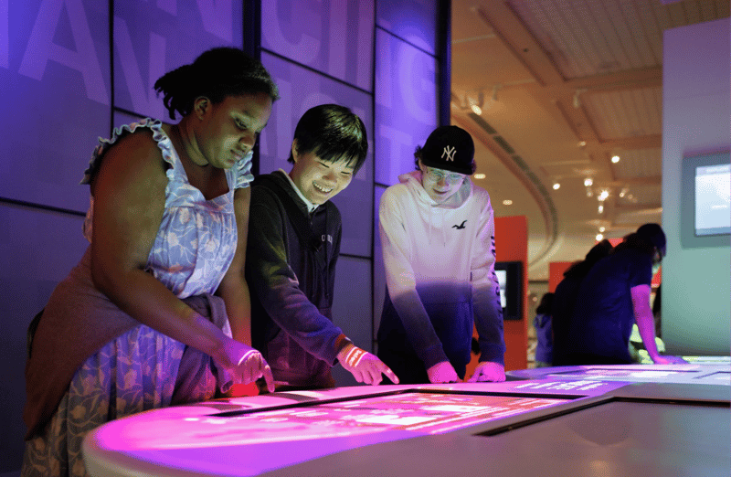 A group of students interact with illuminated touchscreen displays in a futuristic, high-tech learning environment.
