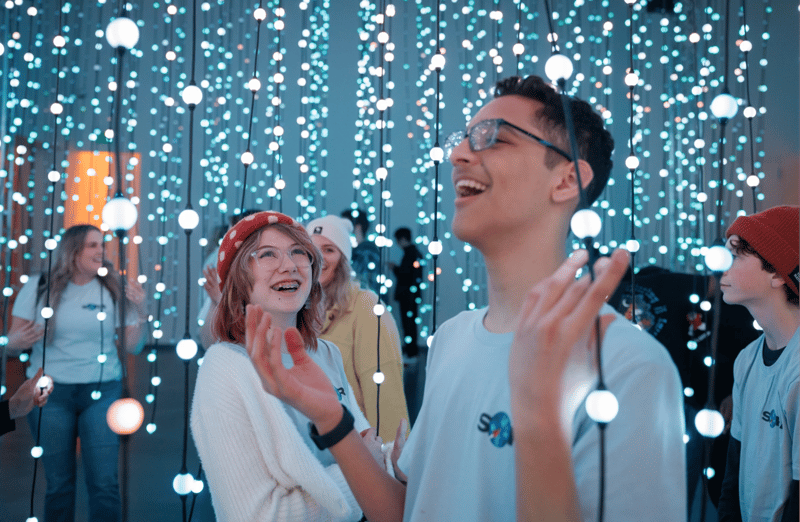 A group of young people wearing matching shirts explore an immersive light installation, smiling and interacting with the glowing displays.