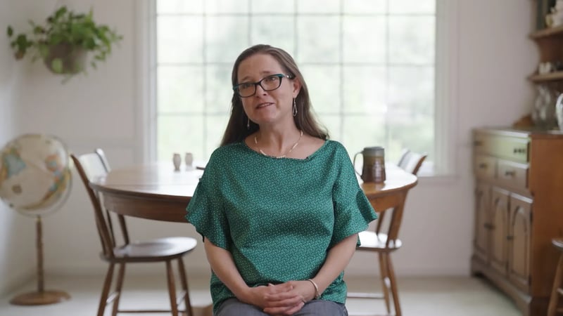 Online homeschool mom wearing a green blouse sits in a well-lit dining room, speaking with a calm and attentive expression.