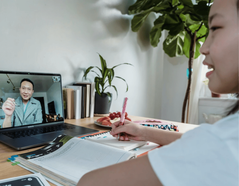 Online homeschool for middle school student engages in a virtual lesson, taking notes while interacting with a teacher on a laptop.